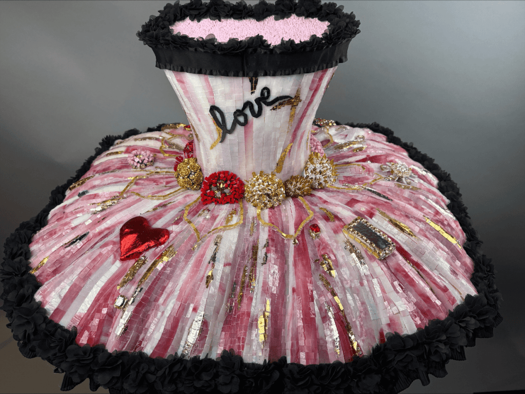 A decorative pink and white dress form, embellished with various ornaments including flowers, a red heart, and gold accents, featuring the word 'love' stitched on the bodice.