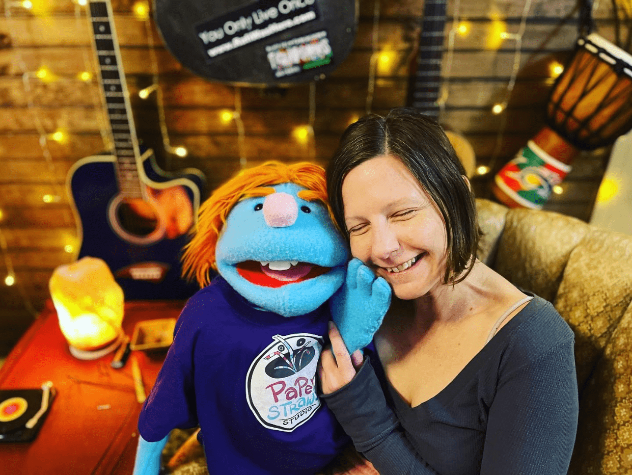 A woman and a blue puppet with orange hair happily pose together in a cozy room with warm lighting and musical instruments in the background.
