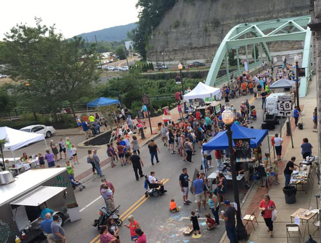 A bustling outdoor event with a large crowd gathered on a street, featuring food trucks, vendor tents, and children playing. A green bridge and rocky hillside are visible in the background.