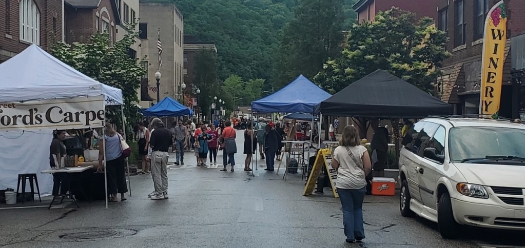 A lively street fair featuring vendor booths with blue and black tents, people walking and browsing, and a sign for a winery in the background.