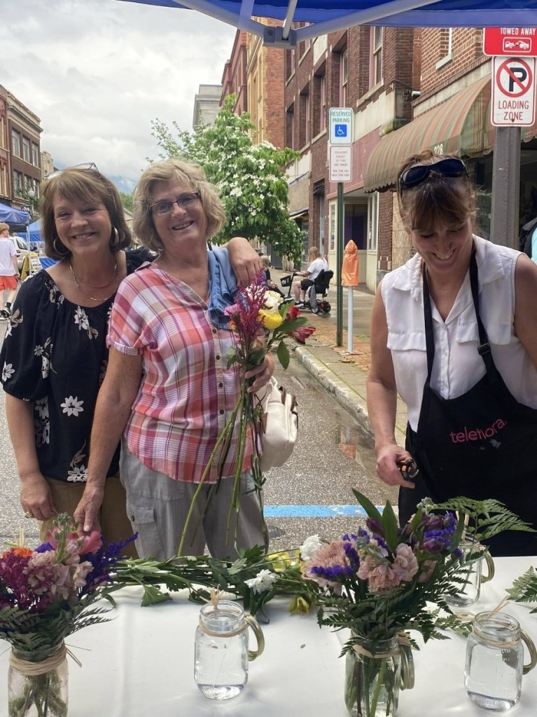 Three women smiling at a market stall, holding flowers, with jars of flowers on the table in front of them. A street scene is visible in the background.