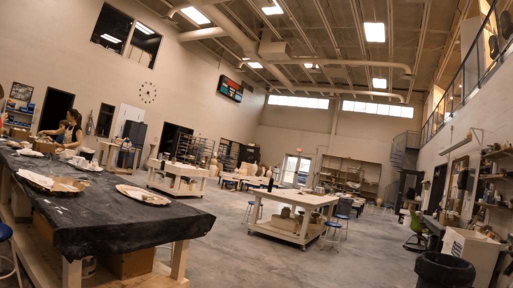 A spacious pottery studio featuring work tables, pottery tools, and shelves with ceramic pieces. Two people are engaged in crafting at a black table, with natural light coming through large windows.
