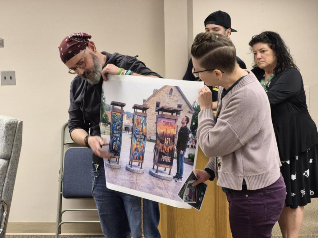 A group of people at a meeting, one man holding a large printed poster showing artistic banners, while a woman examines the poster closely. Two other individuals in the background observe the presentation.