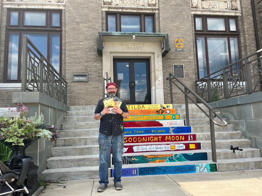 A man wearing a red beanie stands on the steps of a library, holding three children's books. Colorful painted book spines are displayed on the steps, featuring titles like 'Goodnight Moon' and 'Where the Wild Things Are.'
