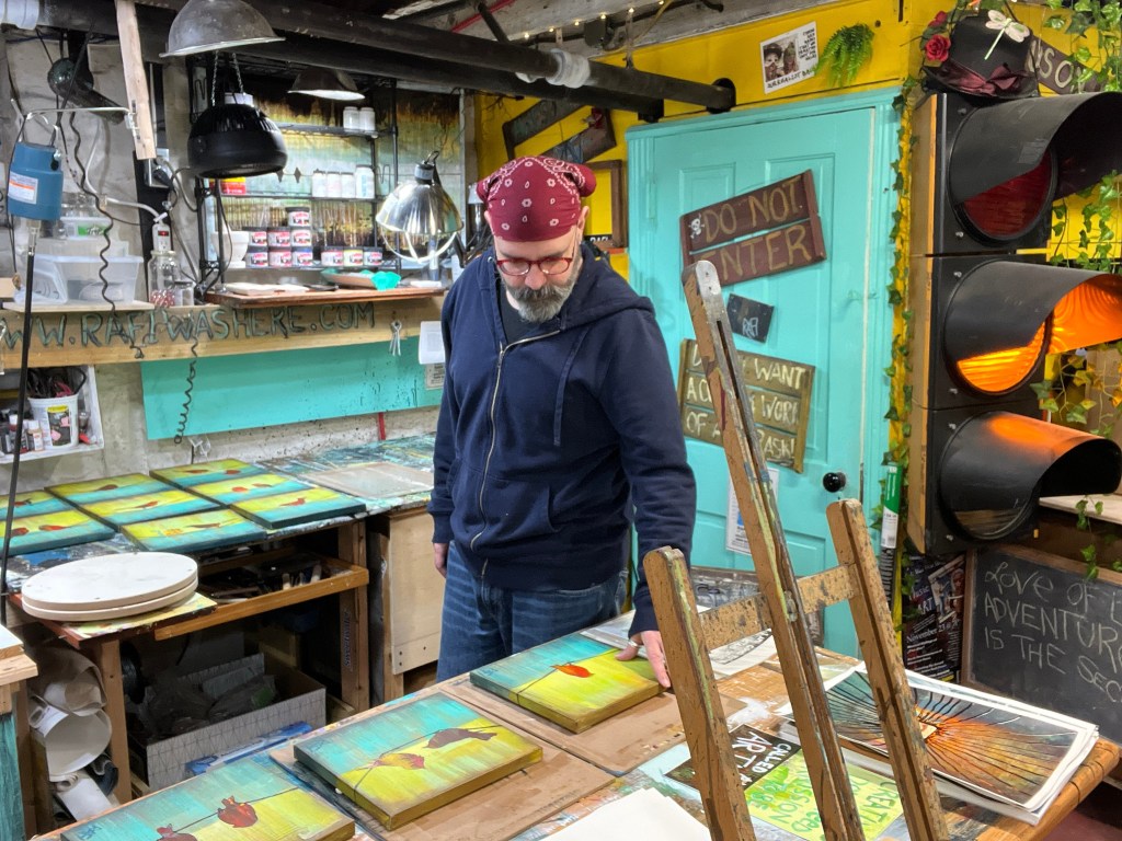An artist in a studio examining colorful paintings on a table, surrounded by art supplies and decorations.
