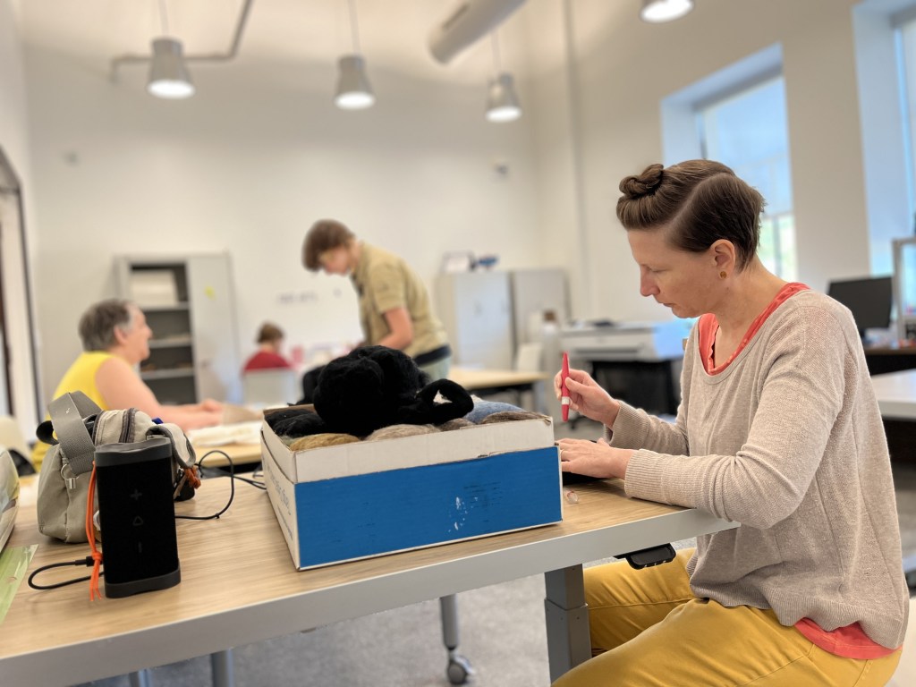 A woman sits at a table, focused on her work while using a red pen. She is surrounded by others engaged in activities in a spacious, well-lit room. Various materials, including a box with colorful items, are visible on the table.