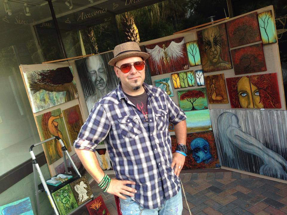 A man wearing a plaid shirt, sunglasses, and a hat stands confidently in front of a display of colorful artwork, featuring various paintings and designs.
