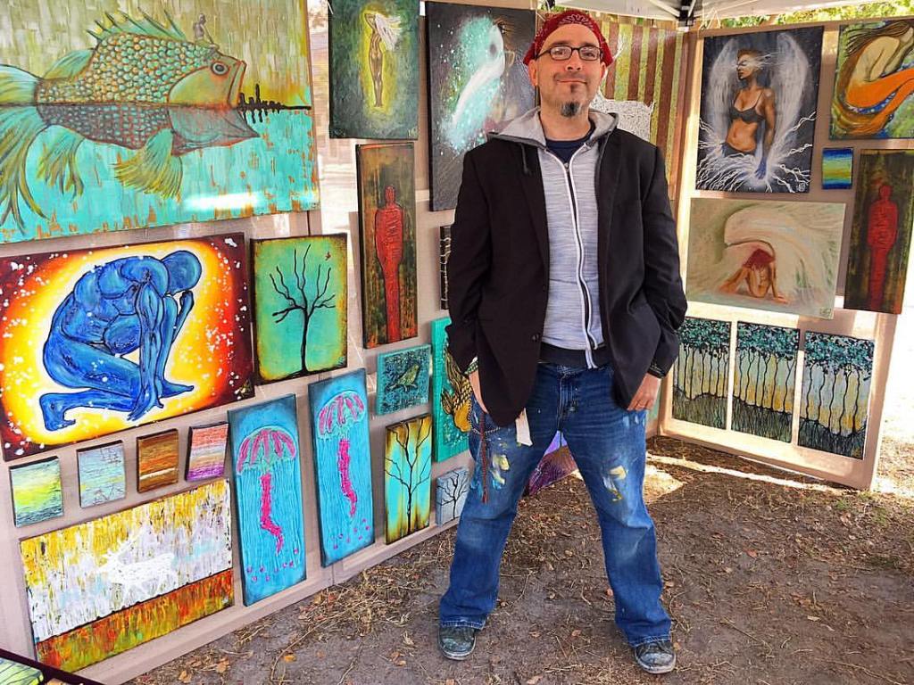 An artist stands confidently in front of a display of colorful paintings, featuring abstract and figurative art, in an outdoor setting.