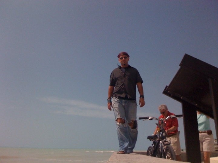 A man with sunglasses and a bandana walks along a pier by the ocean, with a bike and other people in the background.
