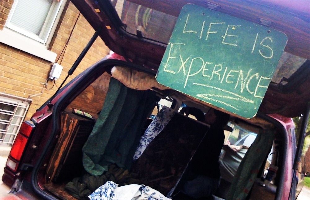 The open trunk of a car displaying a chalkboard sign that reads 'LIFE IS EXPERIENCE' with various items and fabric visible inside.
