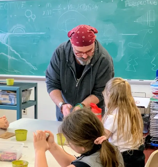 A teacher with a red bandana demonstrates an activity to students at a classroom table, while a chalkboard in the background displays drawings and mathematical equations.