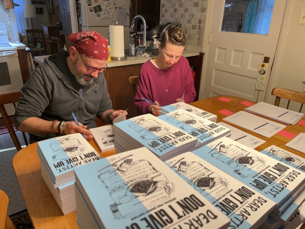 Two individuals signing copies of a book titled 'Dear Artist, Don't Give Up!' at a table filled with stacked books and stationery.