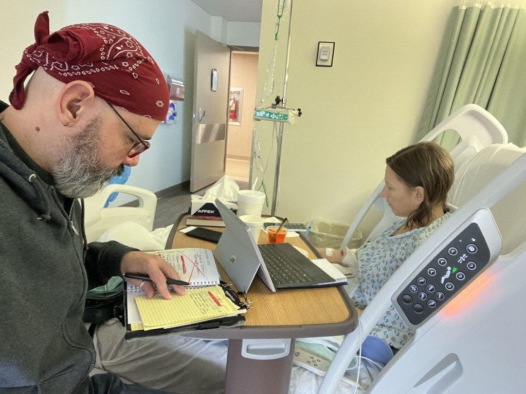A man with a bandana sits at a bedside table in a hospital room, focusing on a notebook, while a woman in a hospital gown uses a laptop. An IV stand is visible nearby, and the room has soft lighting.