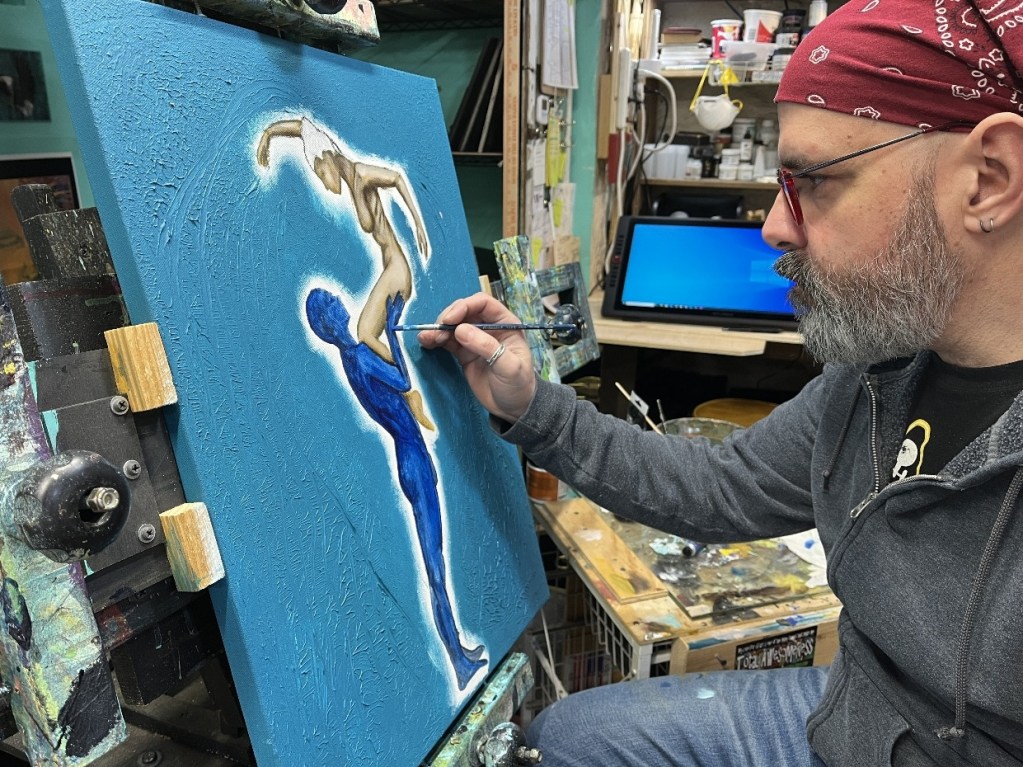 An artist with a beard and glasses is painting on a blue canvas, depicting a figure in dynamic motion, while seated at an easel in a studio.