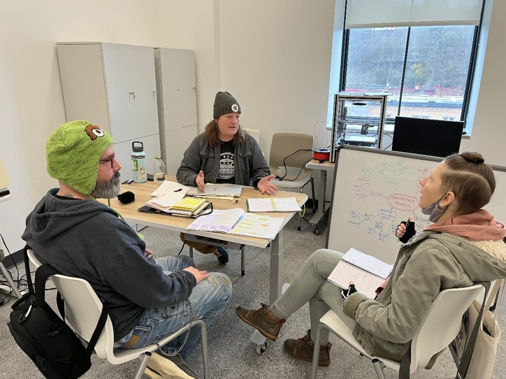 Three individuals engaged in a discussion around a table filled with papers and notebooks in a modern workspace.