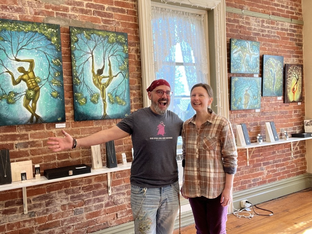 A man and a woman smiling in an art gallery, with colorful paintings on the brick wall behind them and various art pieces displayed on a shelf.