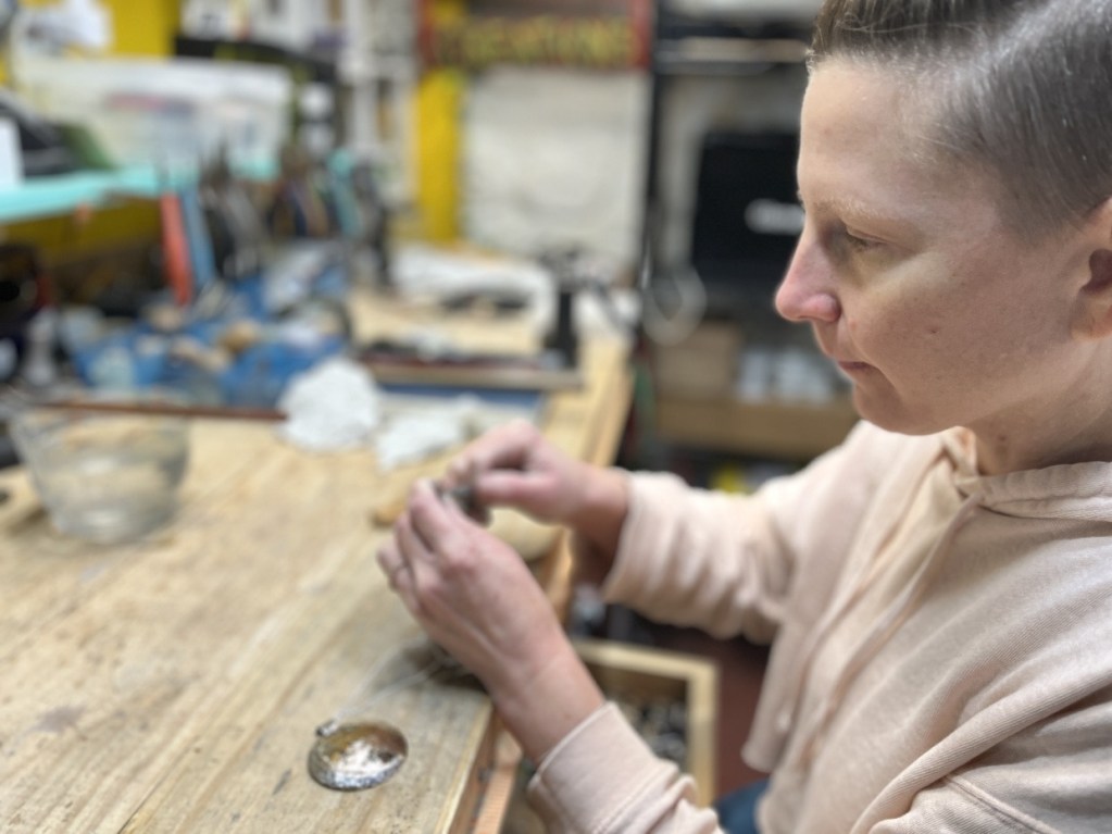 A person working on a craft project at a wooden table, focused on shaping a small object with both hands, surrounded by various tools and materials in a workshop.