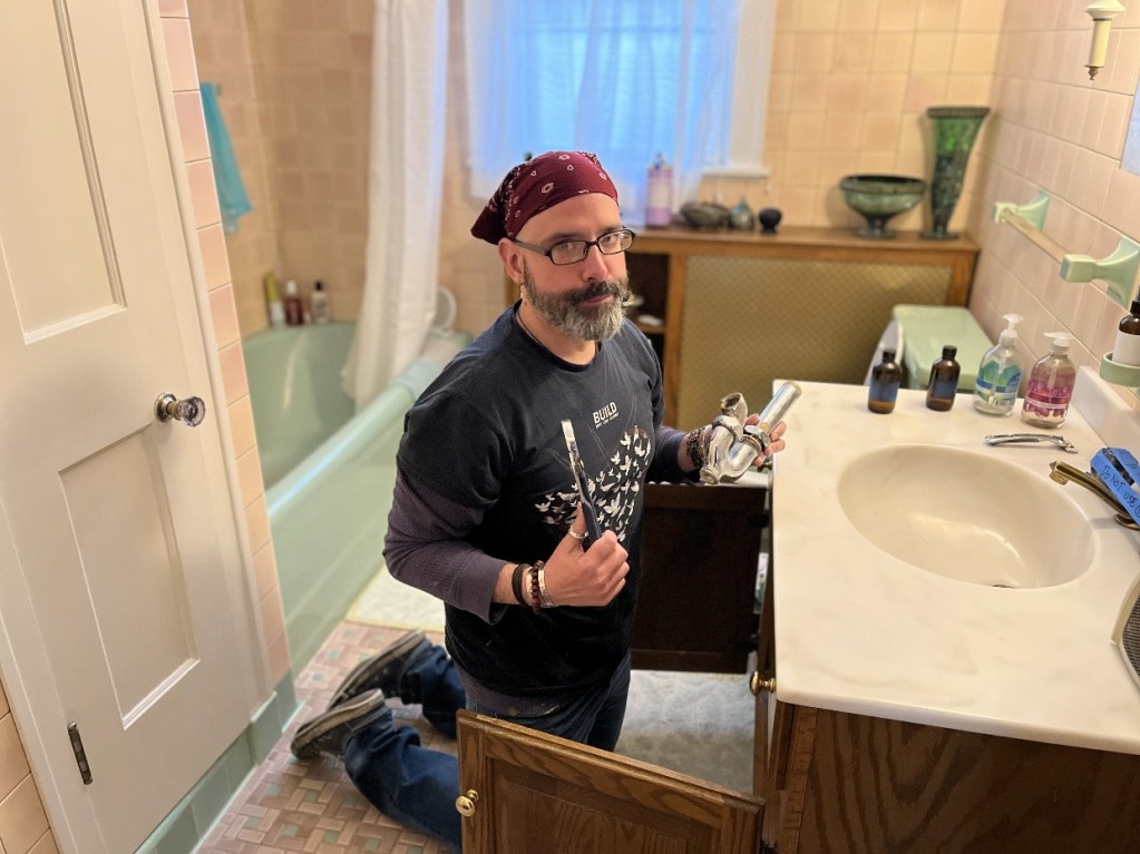 A person kneeling in a bathroom, holding plumbing tools, with an open cabinet and a sink in the foreground.