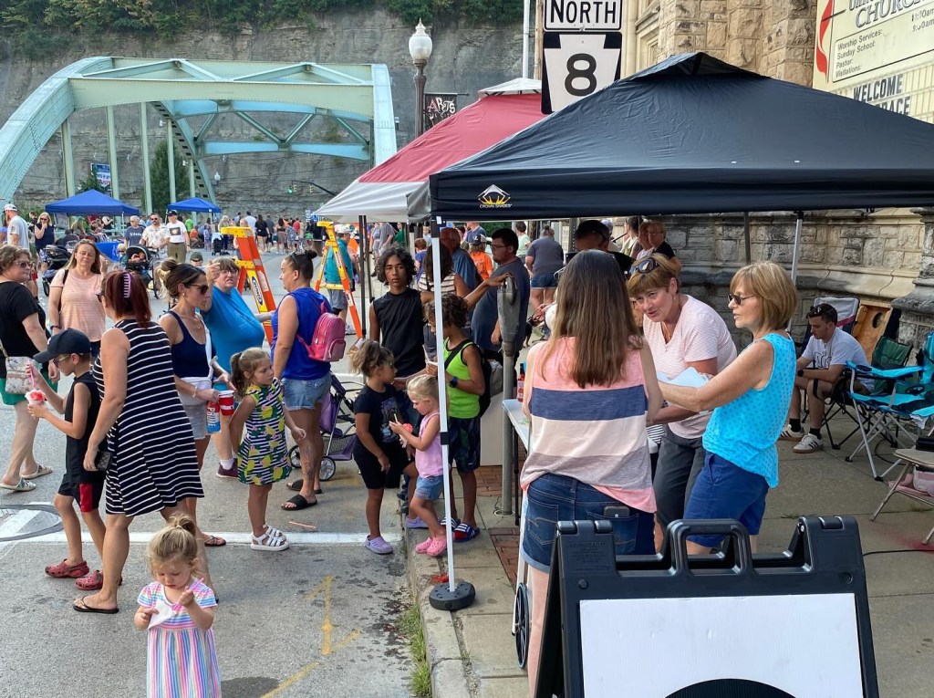 A busy street scene during a community event, featuring various people interacting, children playing, and multiple tents offering food and refreshments. A blue bridge is visible in the background.