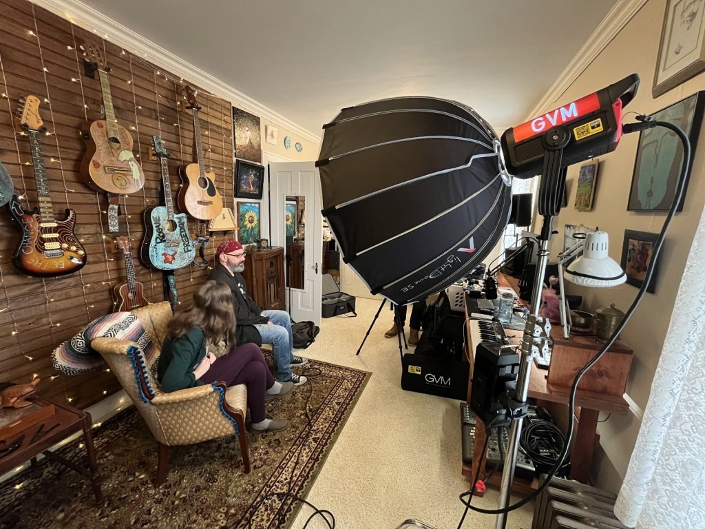 A cozy room with a wall displaying various guitars, featuring two people seated on a vintage couch. One person is wearing a hat and both are looking towards a large softbox light setup, with recording equipment and decorative items in the background.