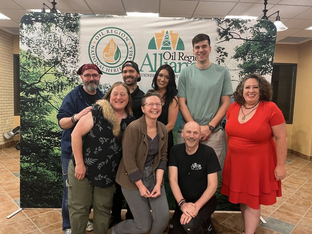 A group of seven individuals posing for a photo in front of a backdrop featuring a logo for the Oil Region National Heritage Area.