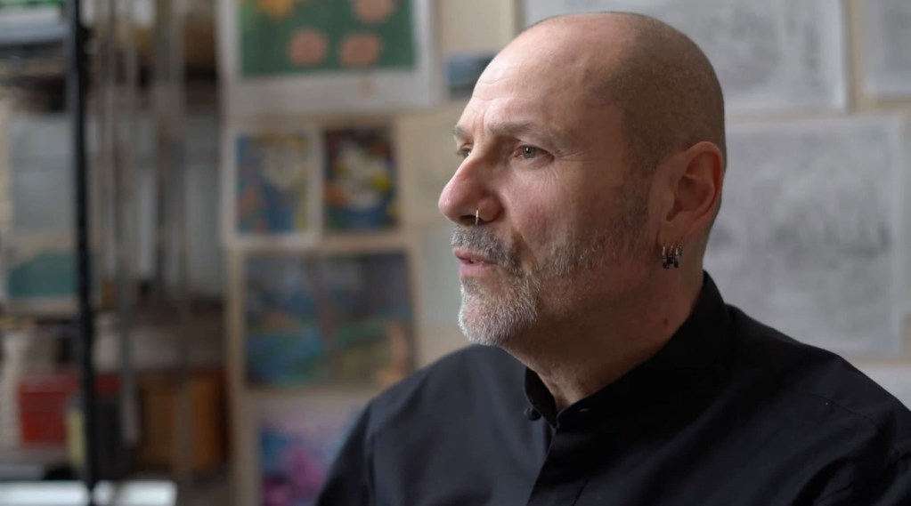 A man with a bald head and earring sits in profile, engaged in conversation, with colorful artwork in the background.