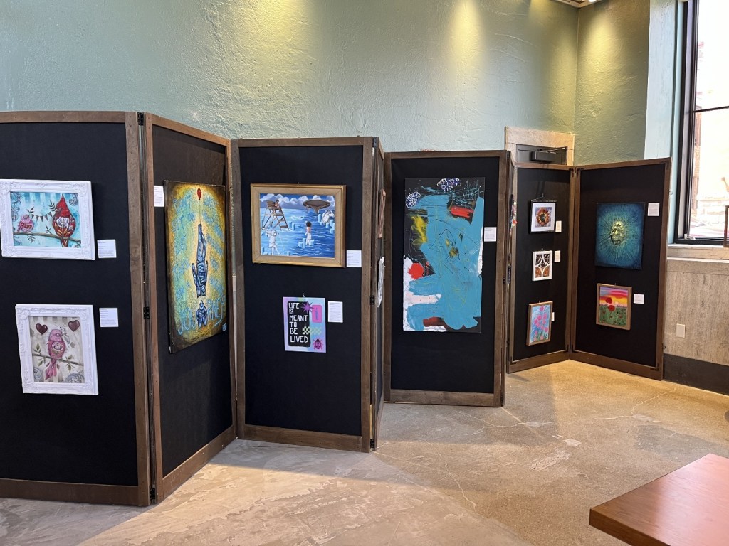 An art exhibition featuring various framed artworks displayed on black partitions in a well-lit gallery setting.