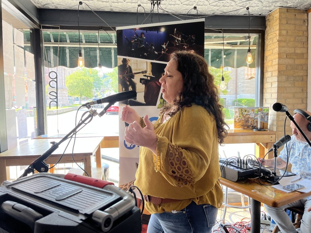 A singer performing at a microphone inside a café, wearing a yellow top with decorative sleeves. In the background, a sound mixing setup and a poster featuring musicians are visible.