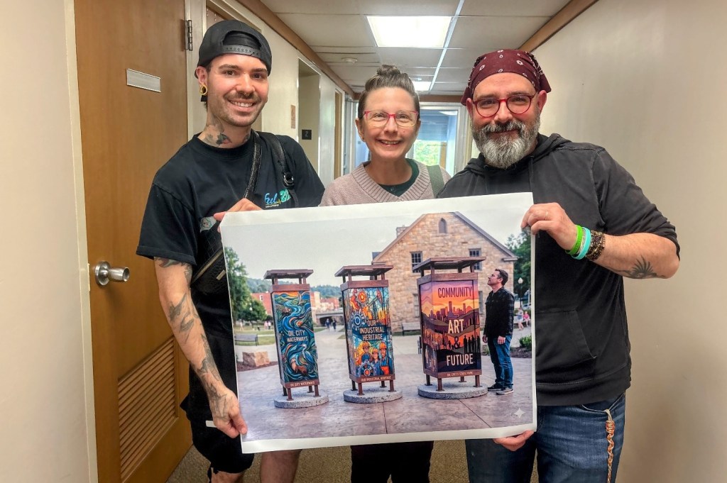 Three individuals pose together in a hallway, holding a large photograph of art installations labeled 'Community Art Future.' The artwork features colorful designs on light poles. The scene is vibrant and reflects a community project.