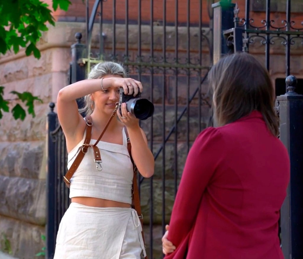 A woman wearing a harness is taking a photo with a camera, while another woman in a red jacket poses nearby.