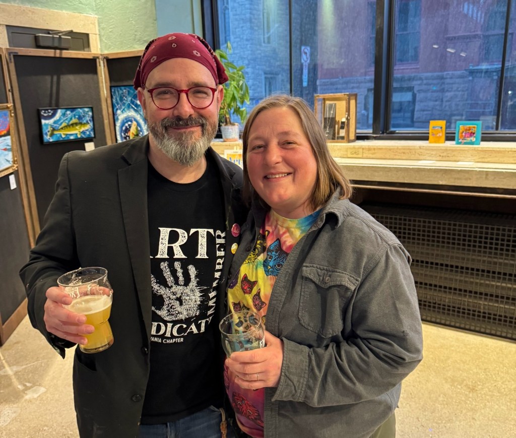 Two smiling individuals holding drinks in a lively setting, with artwork displayed in the background.