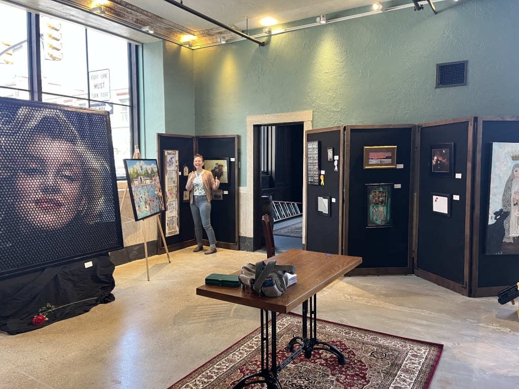 An art exhibition featuring various pieces displayed on black panels, with a person gesturing positively in the foreground. A portrait of a celebrity is prominently featured, alongside other artwork and a small table.