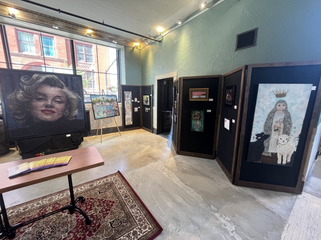 An art gallery featuring various framed artworks. In the foreground, a table with brochures and a rug. Prominent artworks include a large mosaic-style portrait and mixed-media pieces displayed on black panels.