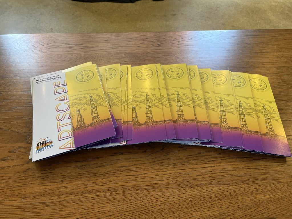A stack of colorful brochures for the Artscape Oil Heritage Festival, featuring illustrations of oil rigs and a vibrant yellow and purple design.