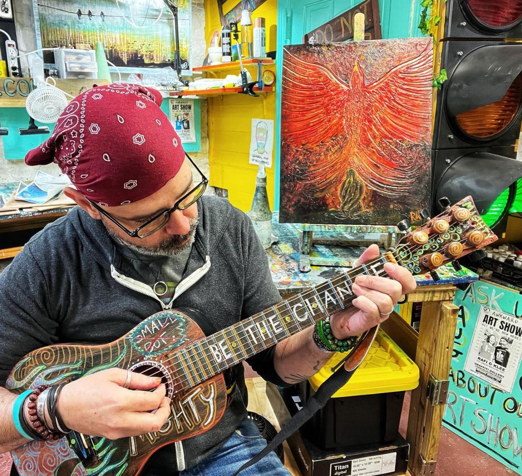 A man with glasses and a bandana plays a colorful, decorated guitar in an artistic workspace filled with various art supplies and a vibrant painting of a phoenix in the background.
