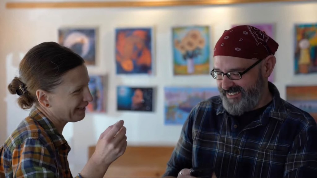 A smiling man with a beard and glasses wearing a red bandana and a plaid shirt, conversing with a woman with a bun hairstyle and a plaid shirt, both looking happy in an art gallery setting with colorful artwork on the walls.