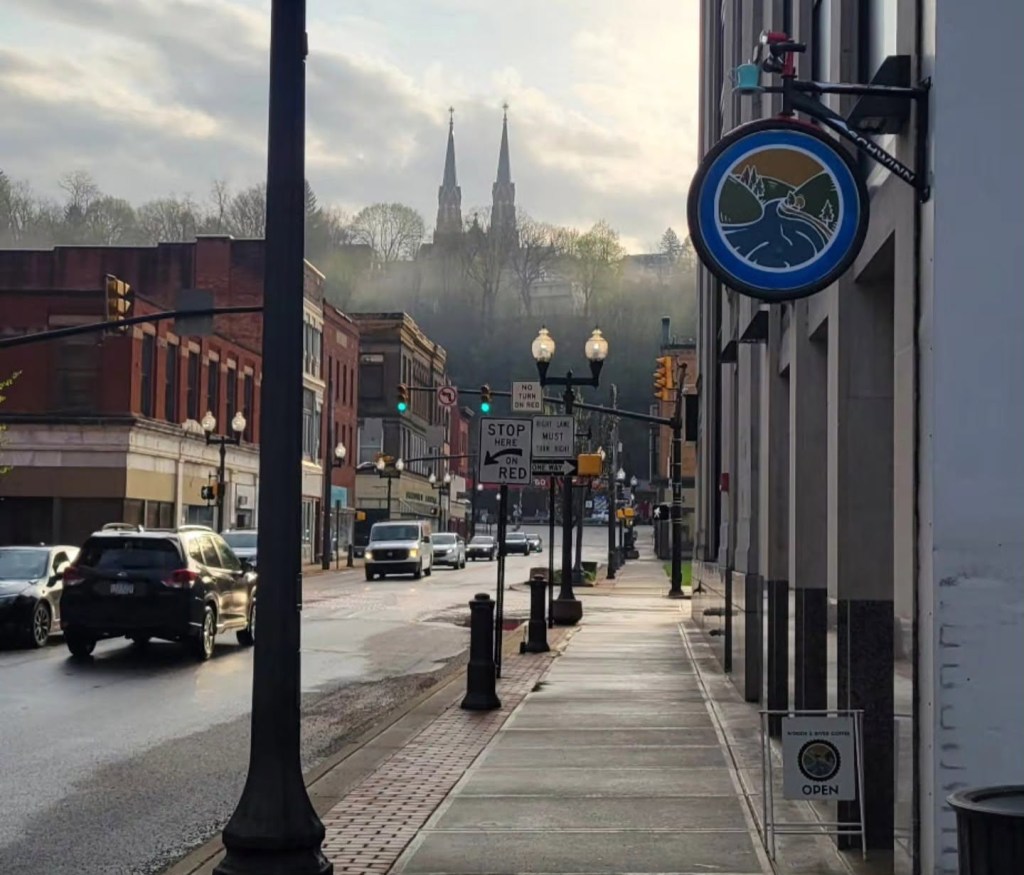 A street view featuring a sidewalk with traffic, vintage buildings, and a prominent church with twin spires in the background. A sign hanging on the building indicates that the business is open.