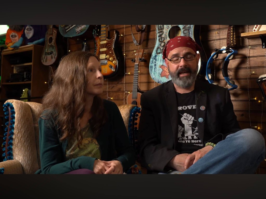 Two people seated in a cozy room filled with musical instruments, including guitars on the wall. One person has long, wavy hair and is wearing a green cardigan, while the other person, wearing a black jacket and a red bandana, is speaking.