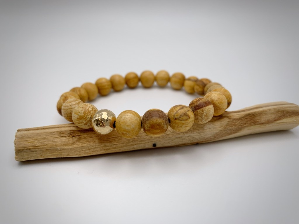 A wooden bead bracelet featuring alternating light and dark wooden beads, with one shiny gold bead, displayed on a piece of driftwood.