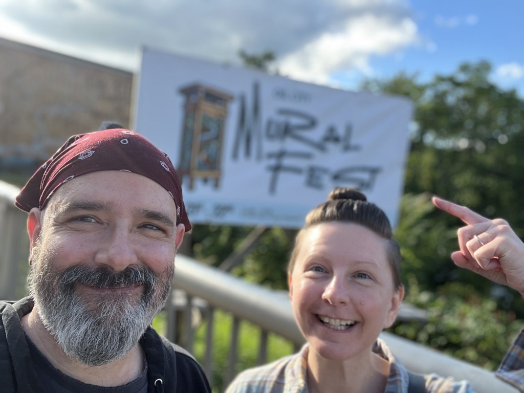 Two people smiling in front of a 'Mural Fest' banner on a sunny day.