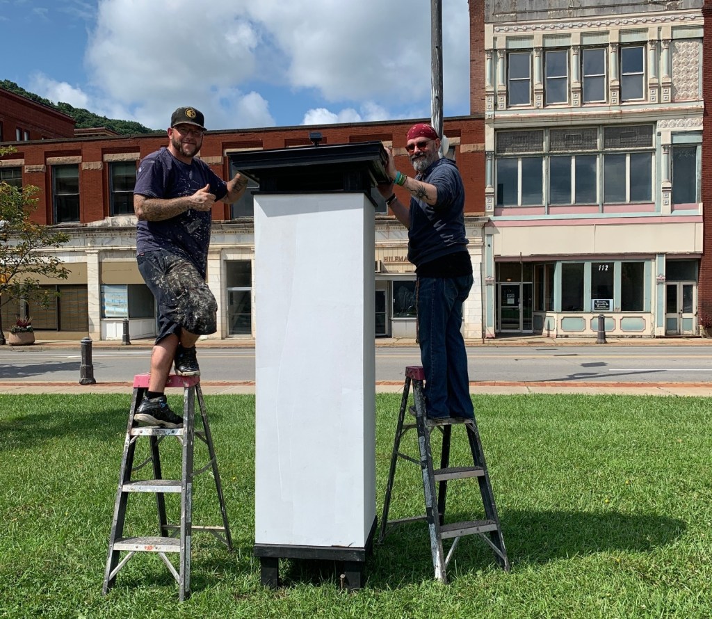 Two men standing on ladders next to a large blank sign structure, smiling and posing for the camera. They are in an outdoor setting with buildings in the background.
