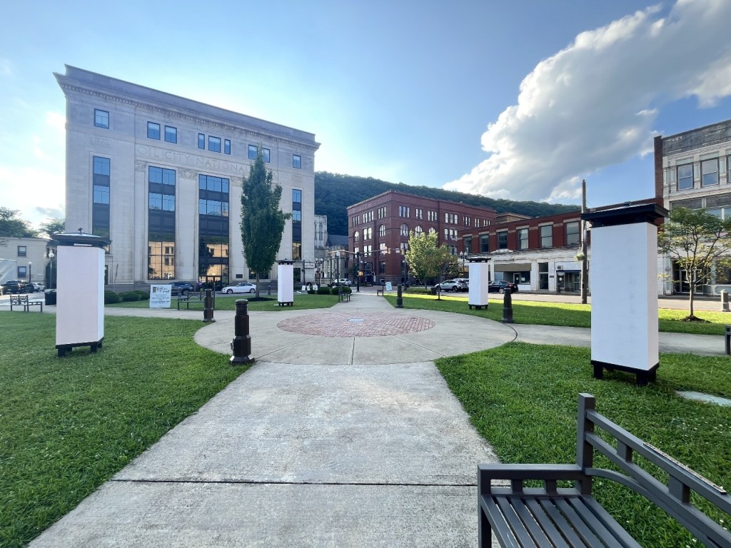A public square featuring green grass, a circular brick design in the center, and several blank display panels. There are benches along the pathways and historic buildings in the background under a blue sky with a few clouds.