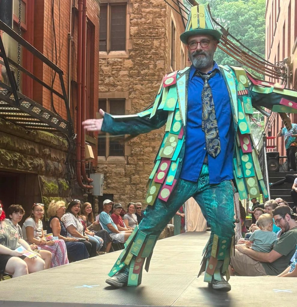 A performer in a colorful, patterned suit with oversized elements walks confidently on a runway in an alley, while an audience observes from the sides.