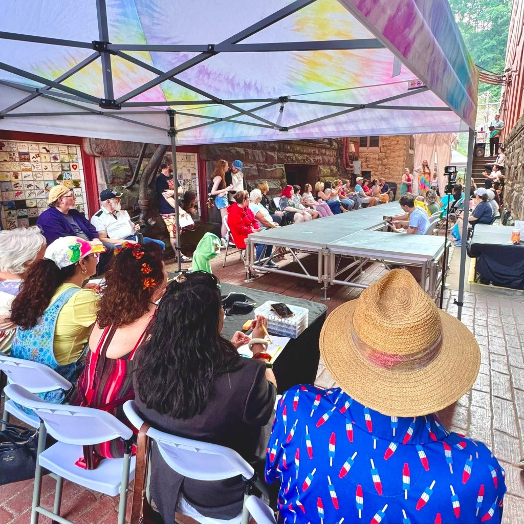A group of people seated under a canopy watching a performance on a stage, with colorful attire and decorations evident among the audience.