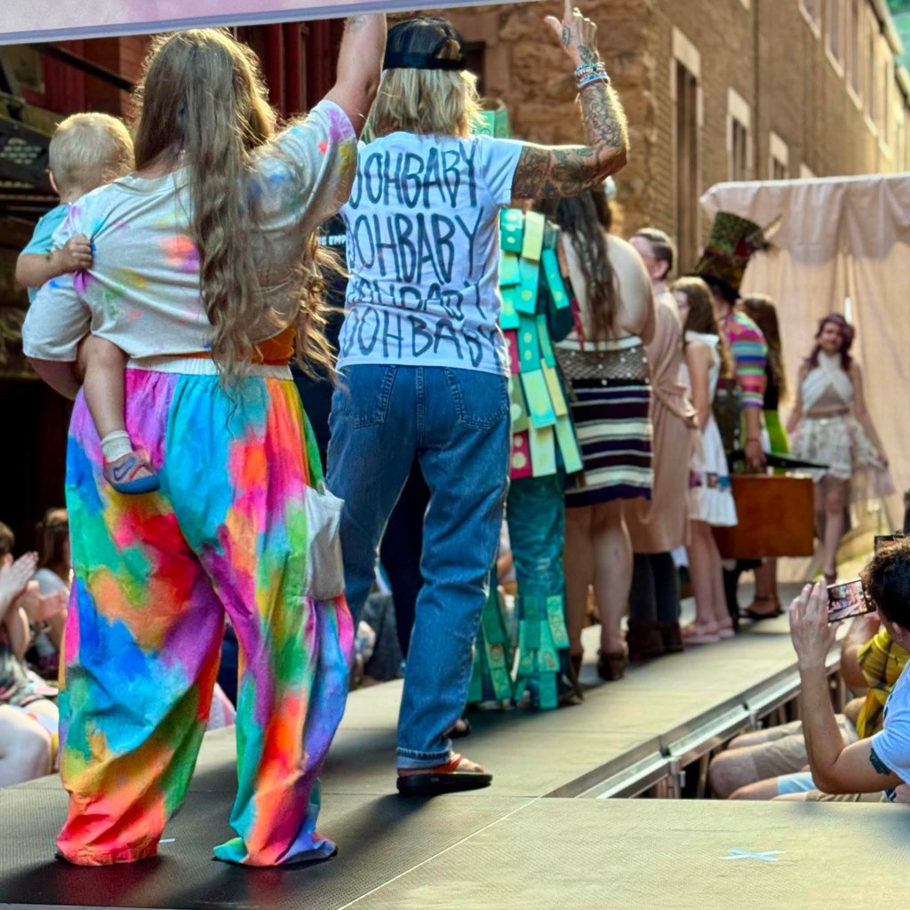 A colorful fashion show scene featuring models walking on a runway, with a woman holding a child on the left and others displaying vibrant outfits in the background.