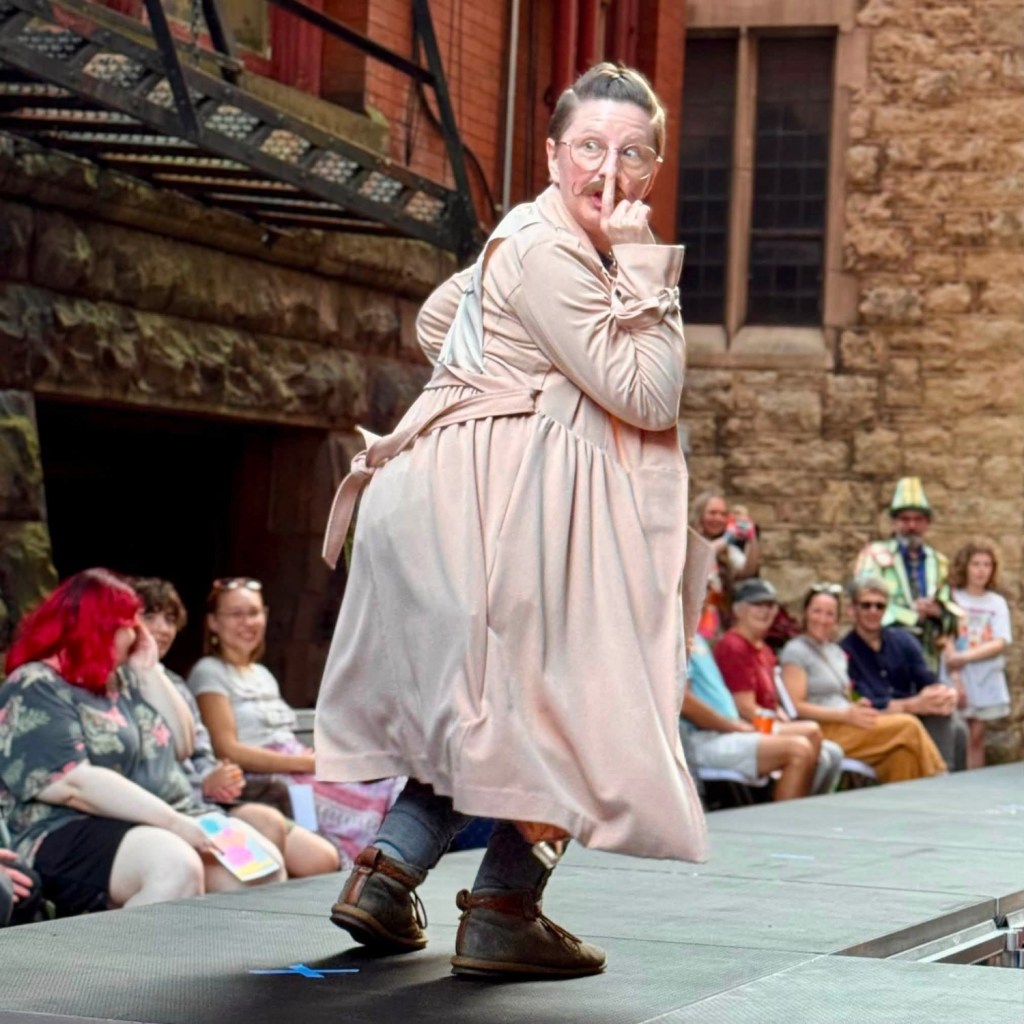 A performer in a light pink oversized dress poses playfully on a runway, holding a finger to their lips in a shushing gesture. The audience, consisting of various individuals, watches with amusement in a lively outdoor setting.