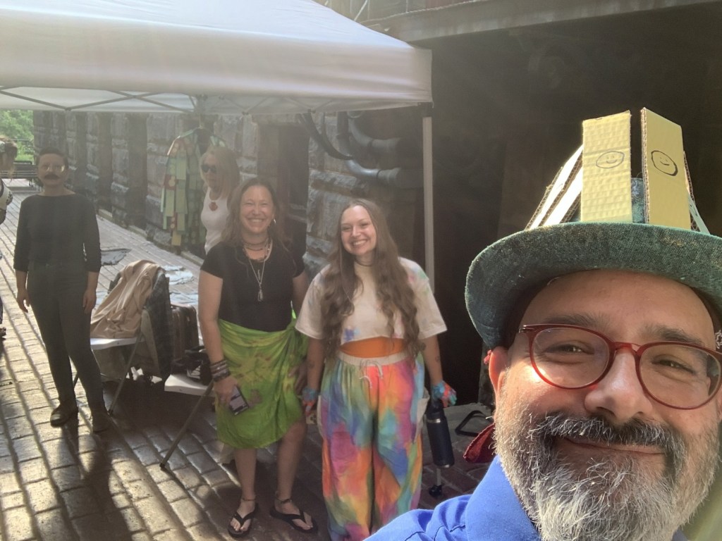 A group of five people posing outdoors in front of a tent, with one person wearing a colorful hat. The background features a stone wall and cobblestone pathway.