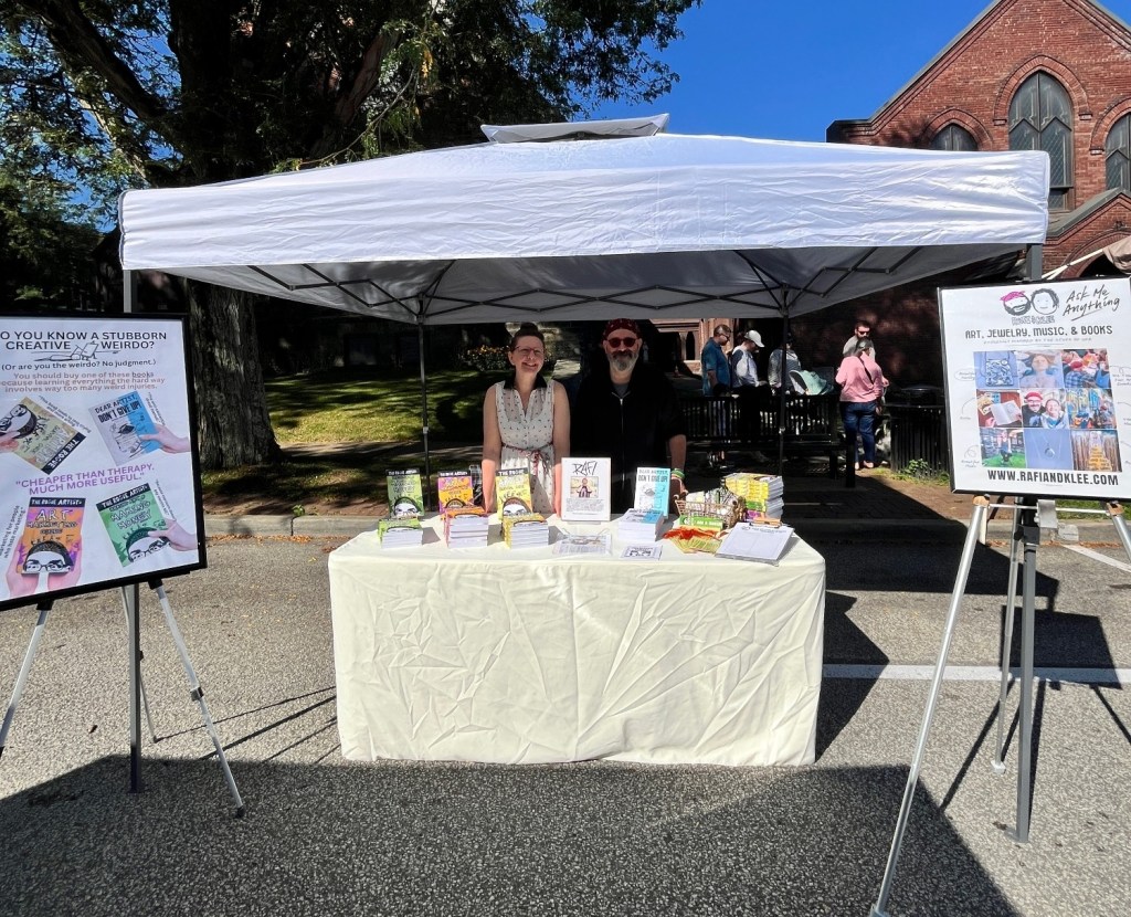 A booth at an outdoor event featuring a couple of vendors with books and merchandise. The table is covered with various creative works, and there's a sign promoting art, jewelry, music, and books. A large banner displays the question 'Do you know a stubborn creative or weirdo?'