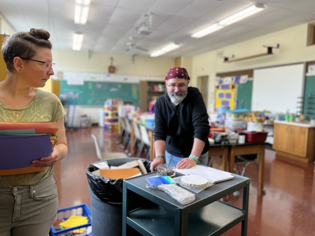 Two adults discussing materials in a classroom, one holding colored paper and the other leaning over a table with craft supplies.