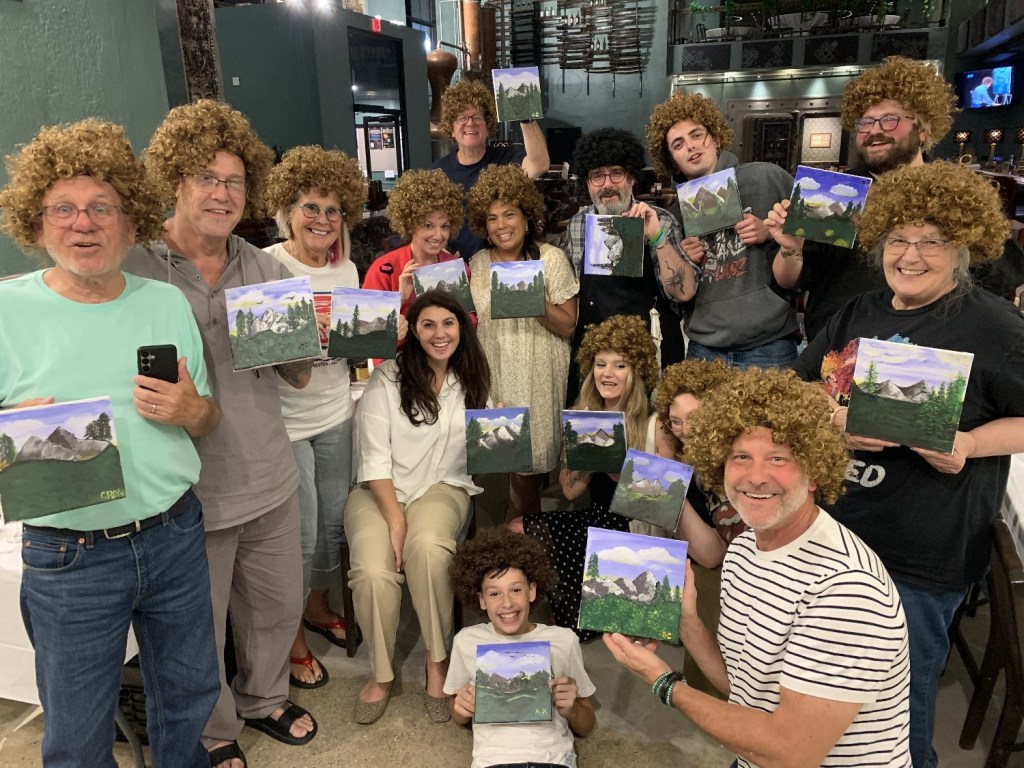 A group of people with curly wigs pose together, each holding a painted canvas featuring mountain landscapes. They are smiling and displaying their artwork in a lively setting.